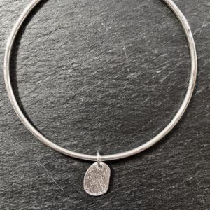 Sterling silver bangle finished with a gentle hammered texture and adorned with a small solid silver beach pebble. The little pebble is cast from a natural beach pebble collected from a beach in Anglesey. Designed and hand made by Carol James of Silverfish Designs.